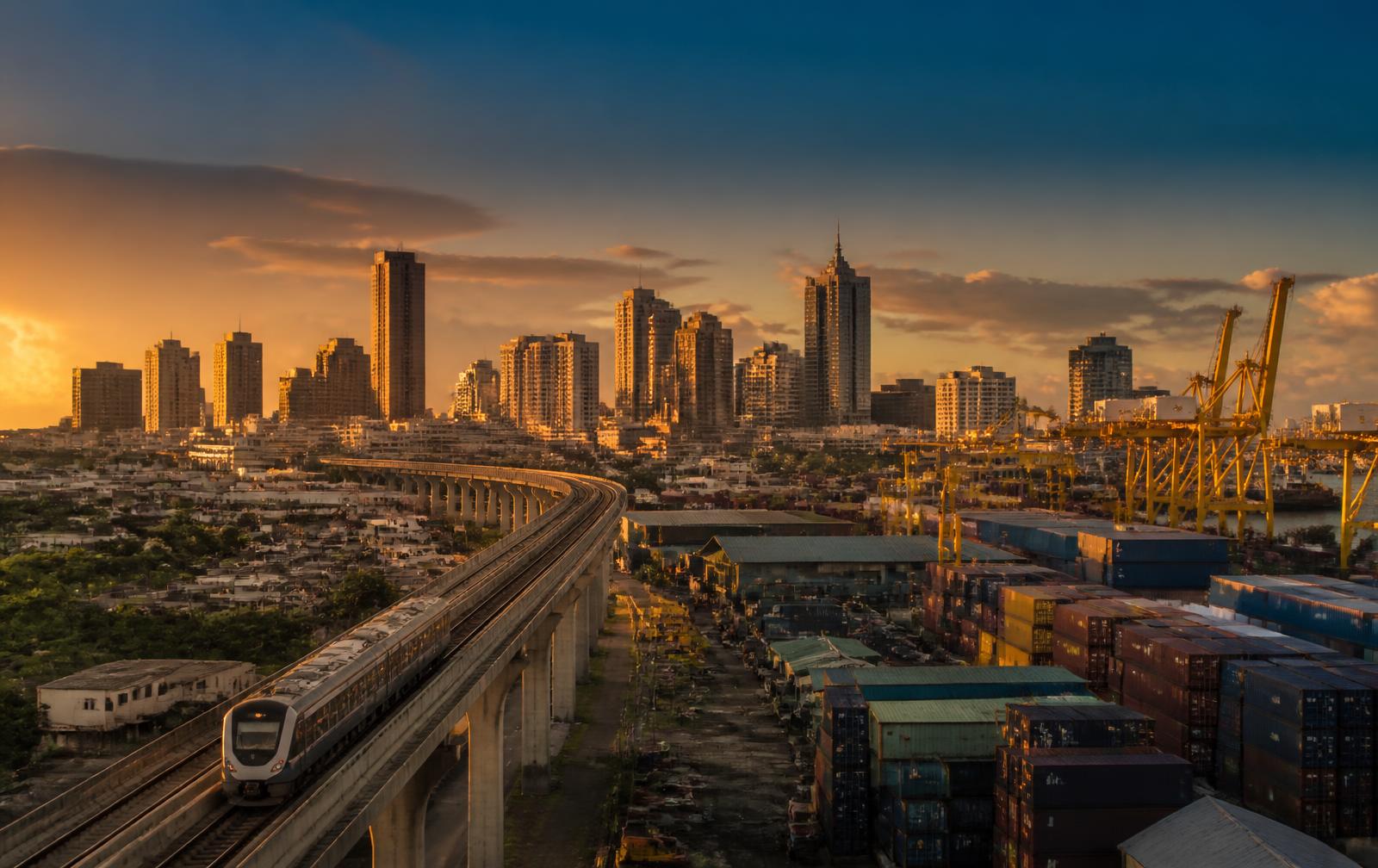 An elevated rail corridor running past a container port and city skyline at golden hour